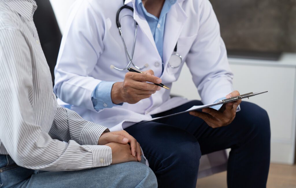 Doctor and patient sitting and talking at medical examination at hospital office, close-up. Therapist filling up medication history records. Medicine and healthcare concept.