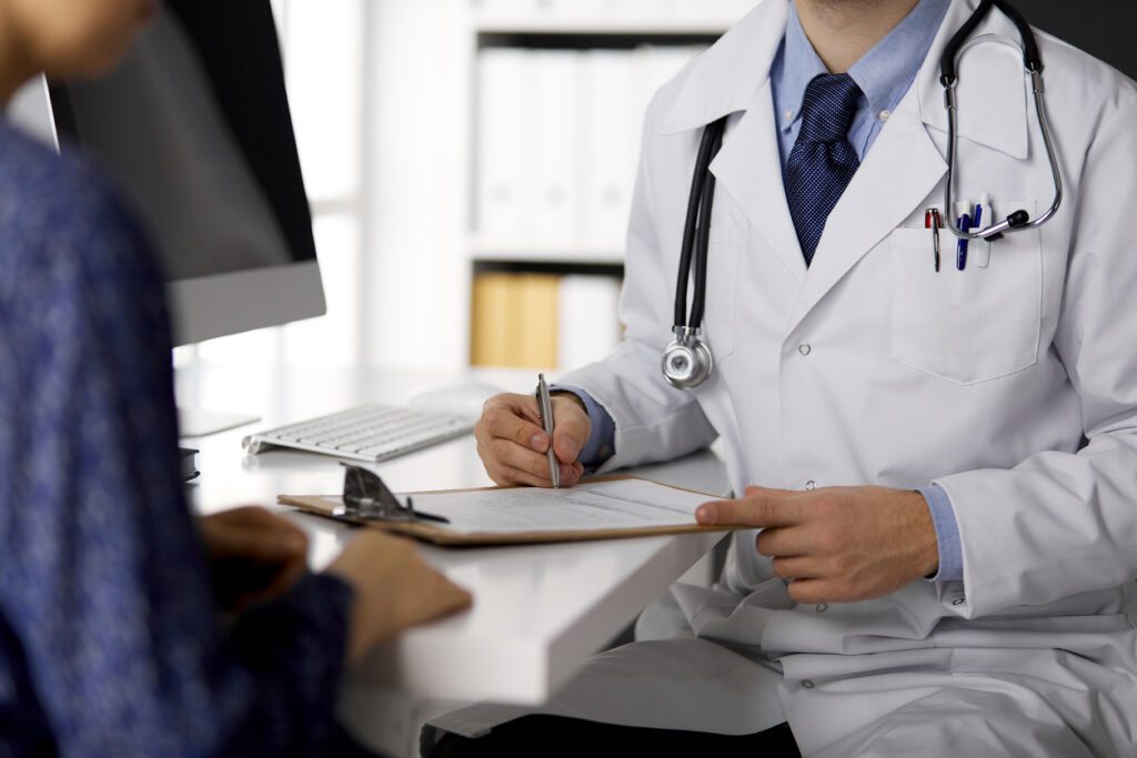 Unknown red-bearded doctor and patient woman discussing current health examination while sitting in sunny clinic, close-up. Medicine concept